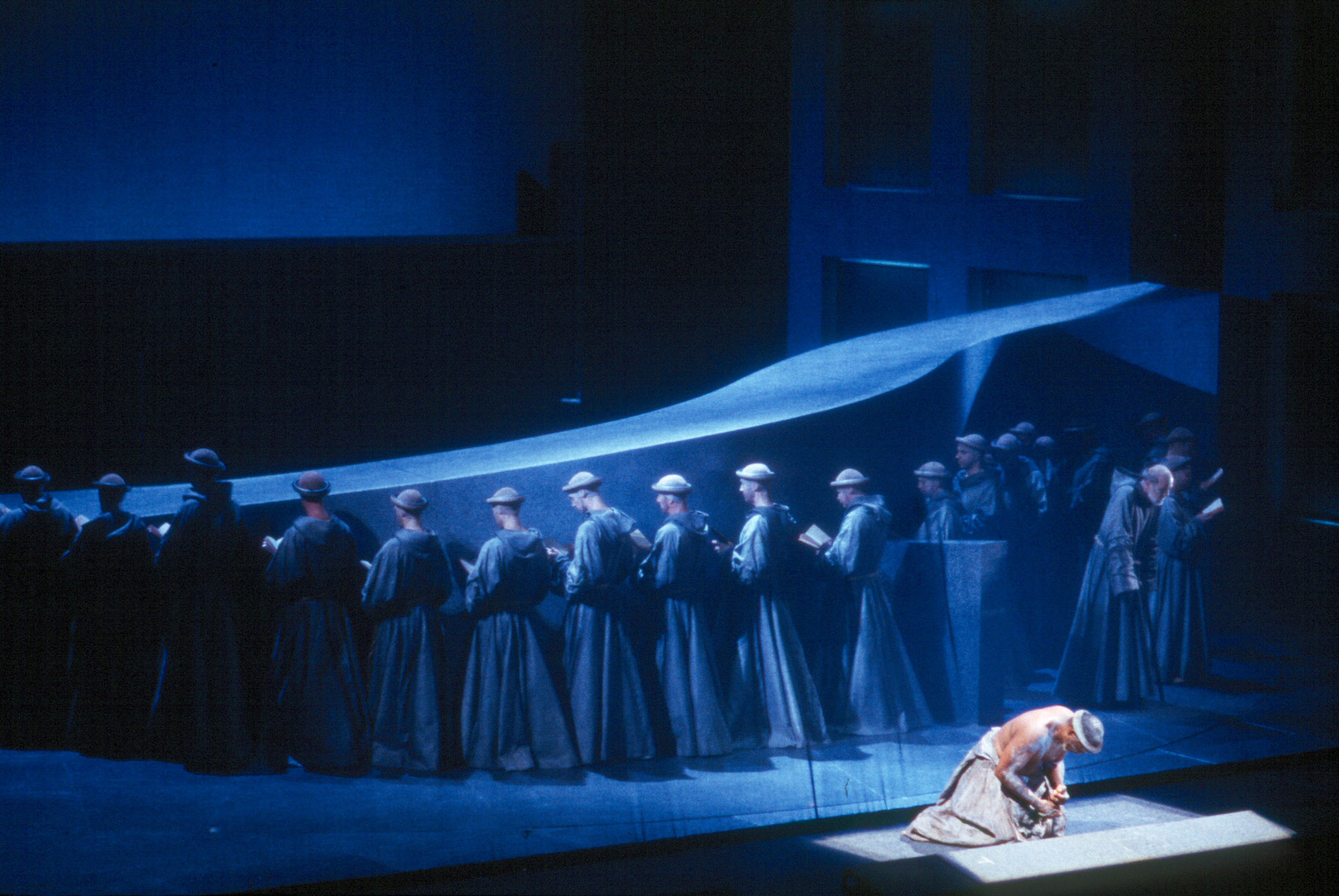 Scene from 2022 SAINT FRANCOIS - monks reading a book with their back turned towards audience as St. Francis is on his knees looking down