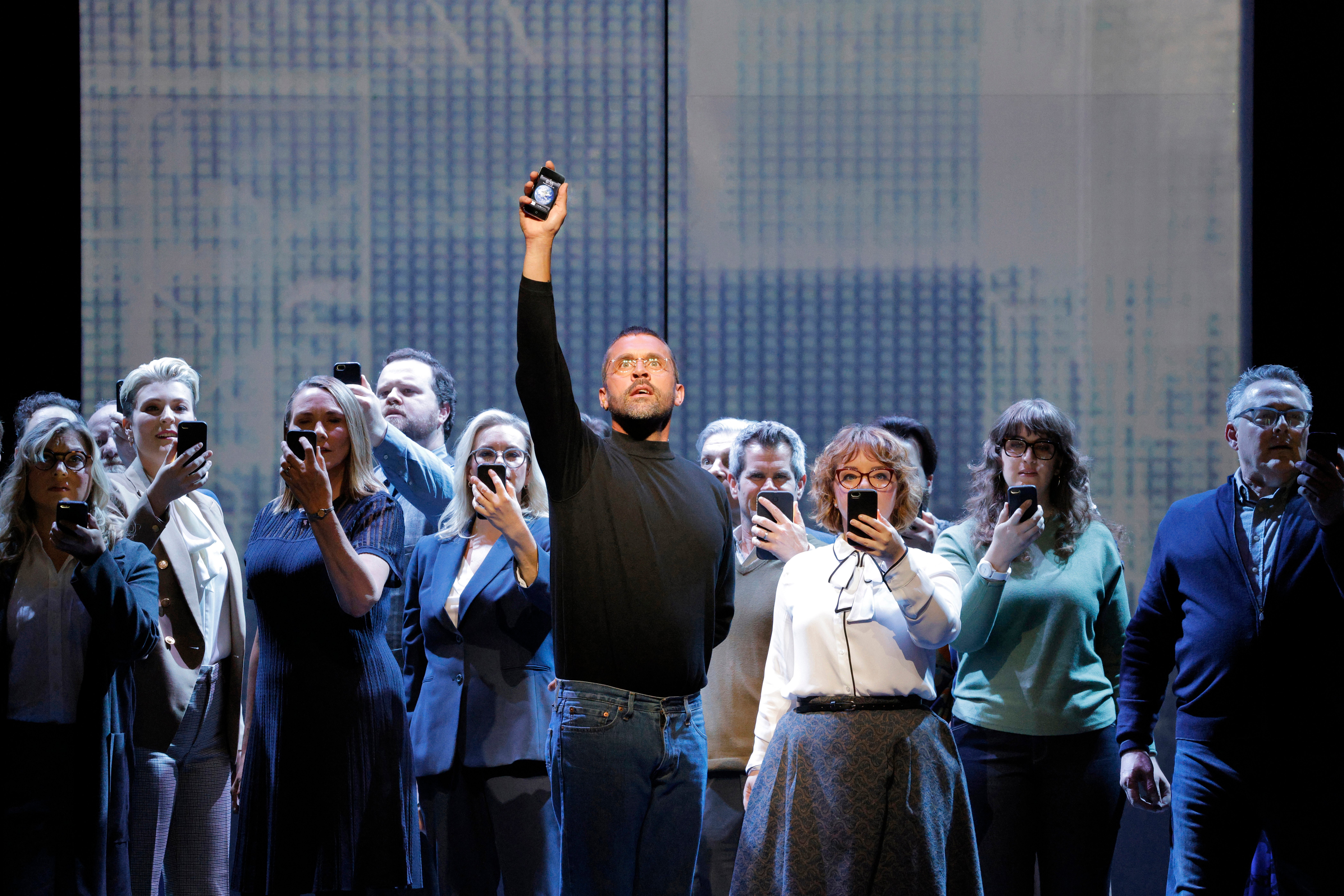 man holding iphone above his head while approximatel 15 other people stand behind him looking at their iphones