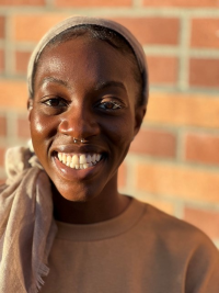 Christabel Nunoo is wearing a tan shirt with a tan headscarf and smiling at the camera.