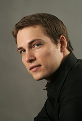 Alek is wearing a dark shirt and smiling at the camera.
